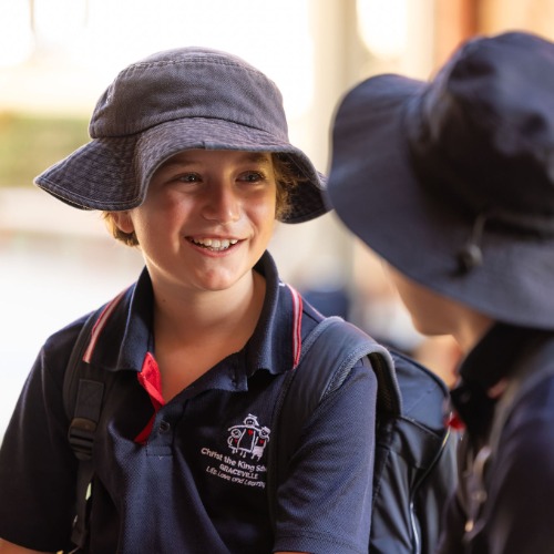 Two students wearing school uniform and hats talking together while carrying backpacks, highlighting friendship, belonging and positive relationships within the primary school environment